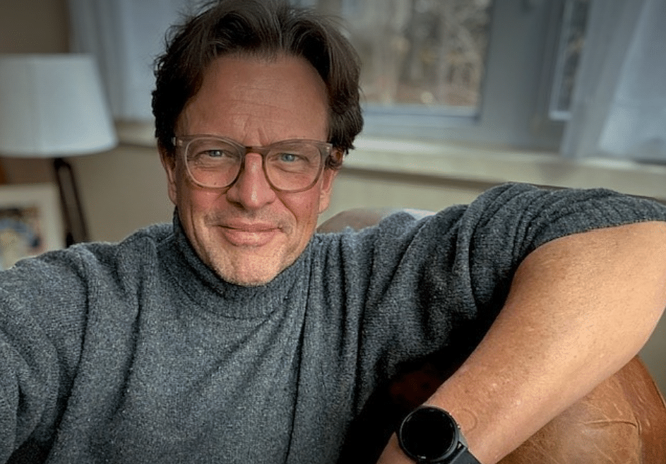 Jay sitting on a leather chair in a cozy room, wearing glasses and a grey sweater, smiling at the camera